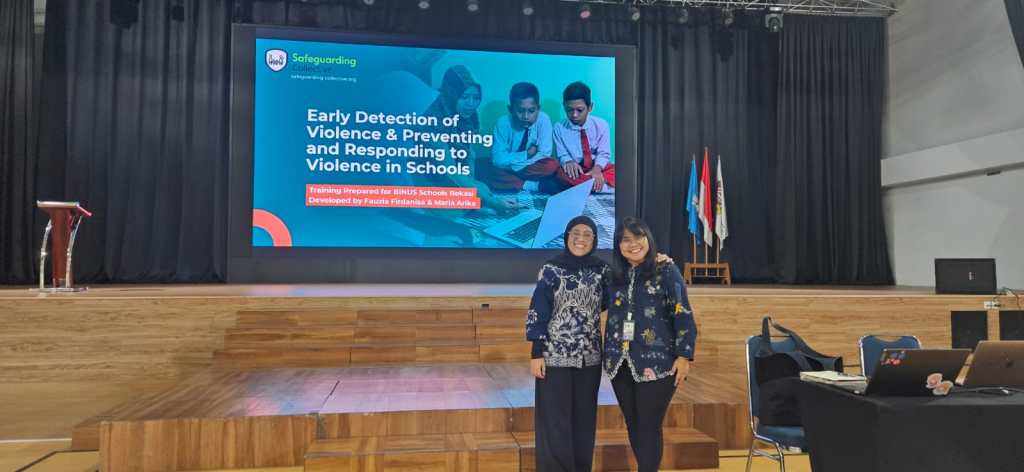 An image of Fauzia and Arika smiling in front of the stage. Behind them, the title slide Ëarly Detection of Violence & Preventing and Responding to Violence in Schools is projected.